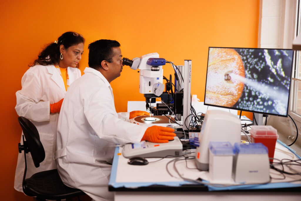 Professors Hameeda Sultana and Girish Neelakanta, wearing white lab coats and orange gloves, look at a tick under a microscope