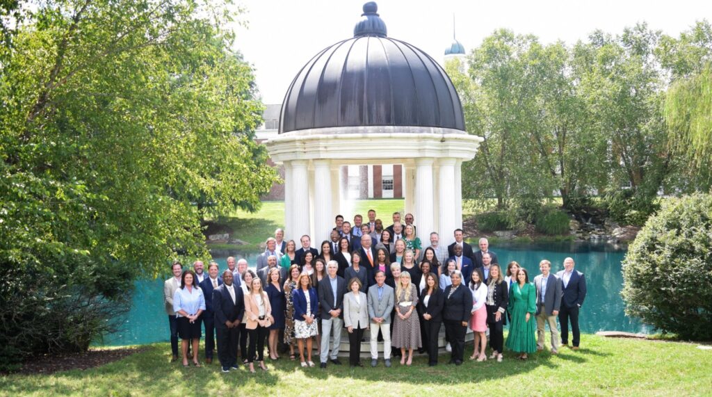 A group of about 50 people stand in front of an outside gazebo with a pond in the background. David Anderson is standing in the middle of the group wearing an orange tie.