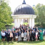 A group of about 50 people stand in front of an outside gazebo with a pond in the background. David Anderson is standing in the middle of the group wearing an orange tie.