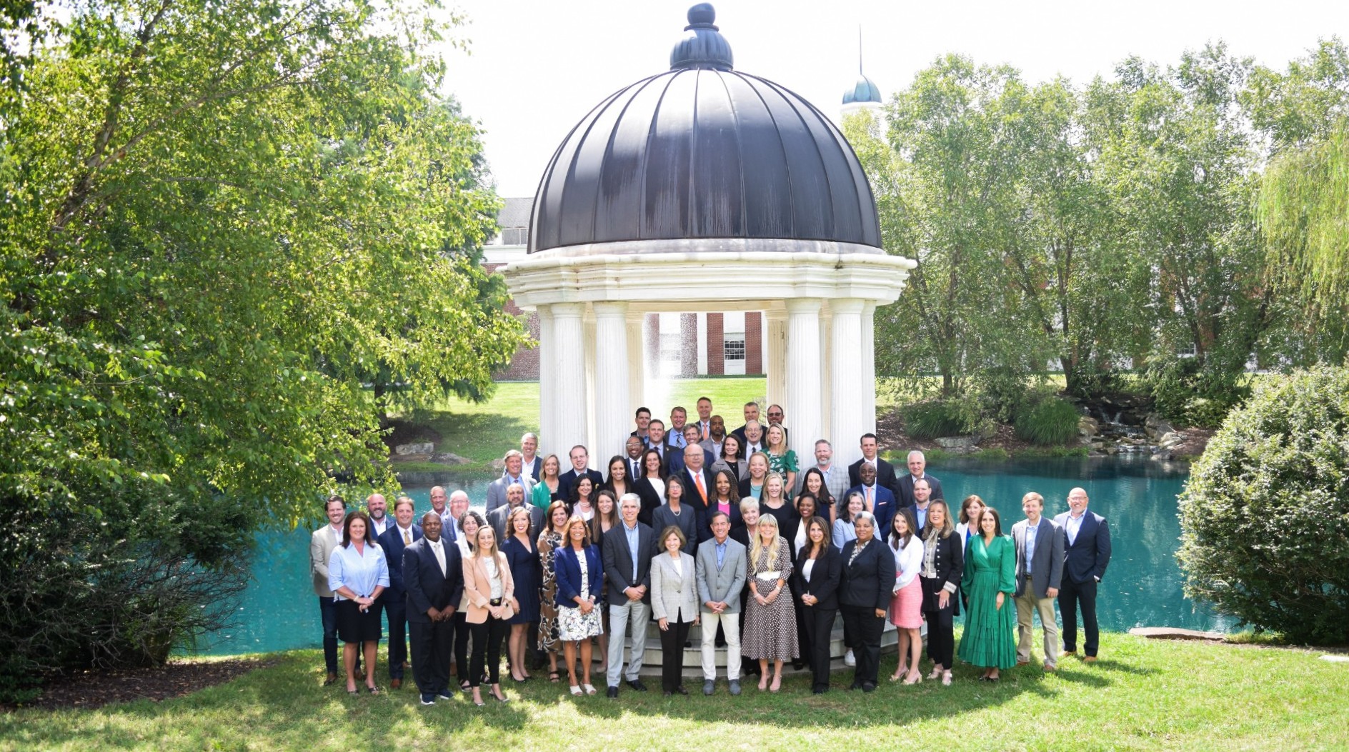 A group of about 50 people stand in front of an outside gazebo with a pond in the background. David Anderson is standing in the middle of the group wearing an orange tie.