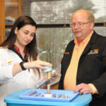 A female graduate student with dark hair in a white lab coat shows a bone model to a man in a black jacket and orange and white checked shirt