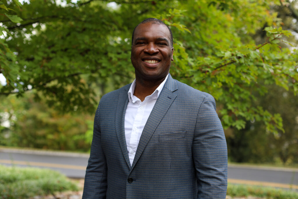 Chika Okafor poses for a headshot wearing a blazer and tie in front of greenery