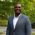 Chika Okafor poses for a headshot wearing a blazer and tie in front of greenery
