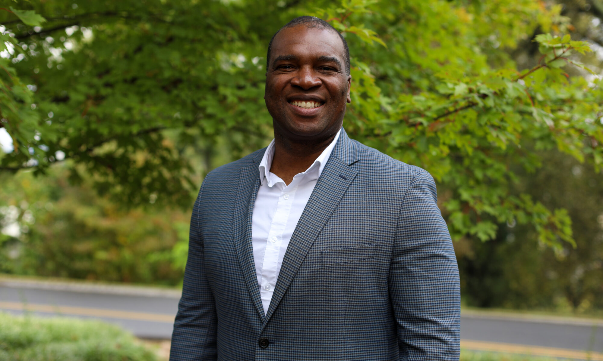 Chika Okafor poses for a headshot wearing a blazer and tie in front of greenery
