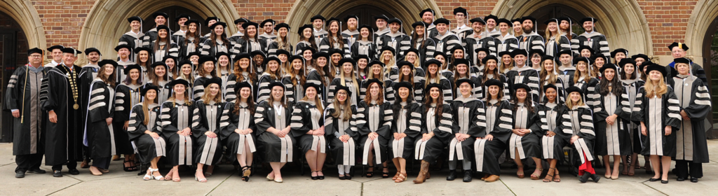 Image of 90 veterinary students and administrators dressed in graduation regalia standing on risers in front of Alumni Memorial Building, a brick building.