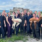 Veterinary students dressed in blue coveralls hold a white and a brown cria in front of a farm house