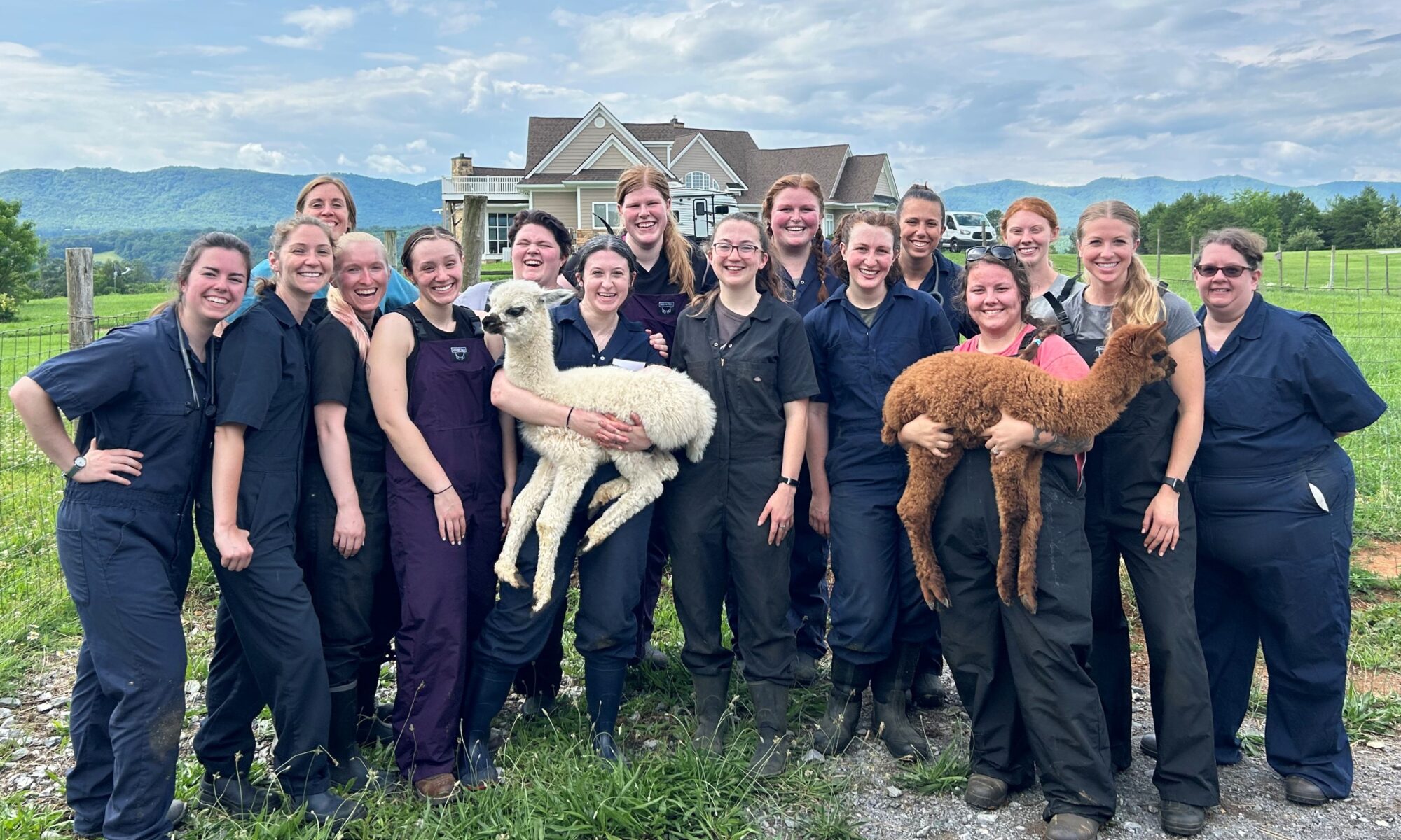 Veterinary students dressed in blue coveralls hold a white and a brown cria in front of a farm house