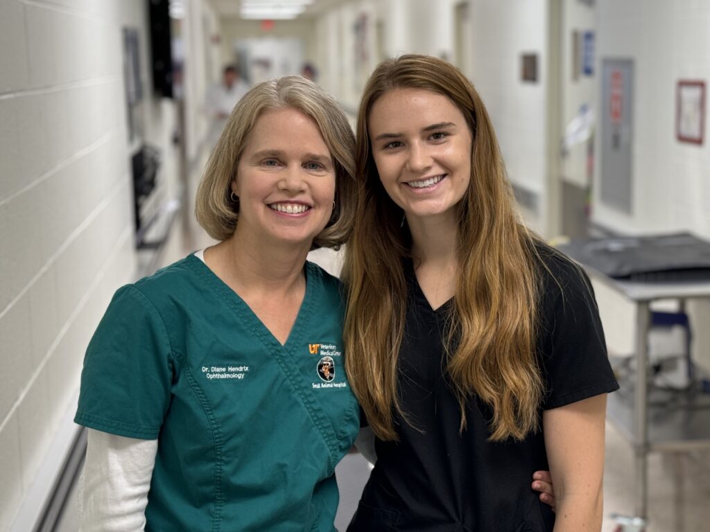 Dr. Diane Hendrix in screen scrubs stands next to her daughter, Emma, who is wearing a black scrub top. They are in a veterinary hospital hallway.