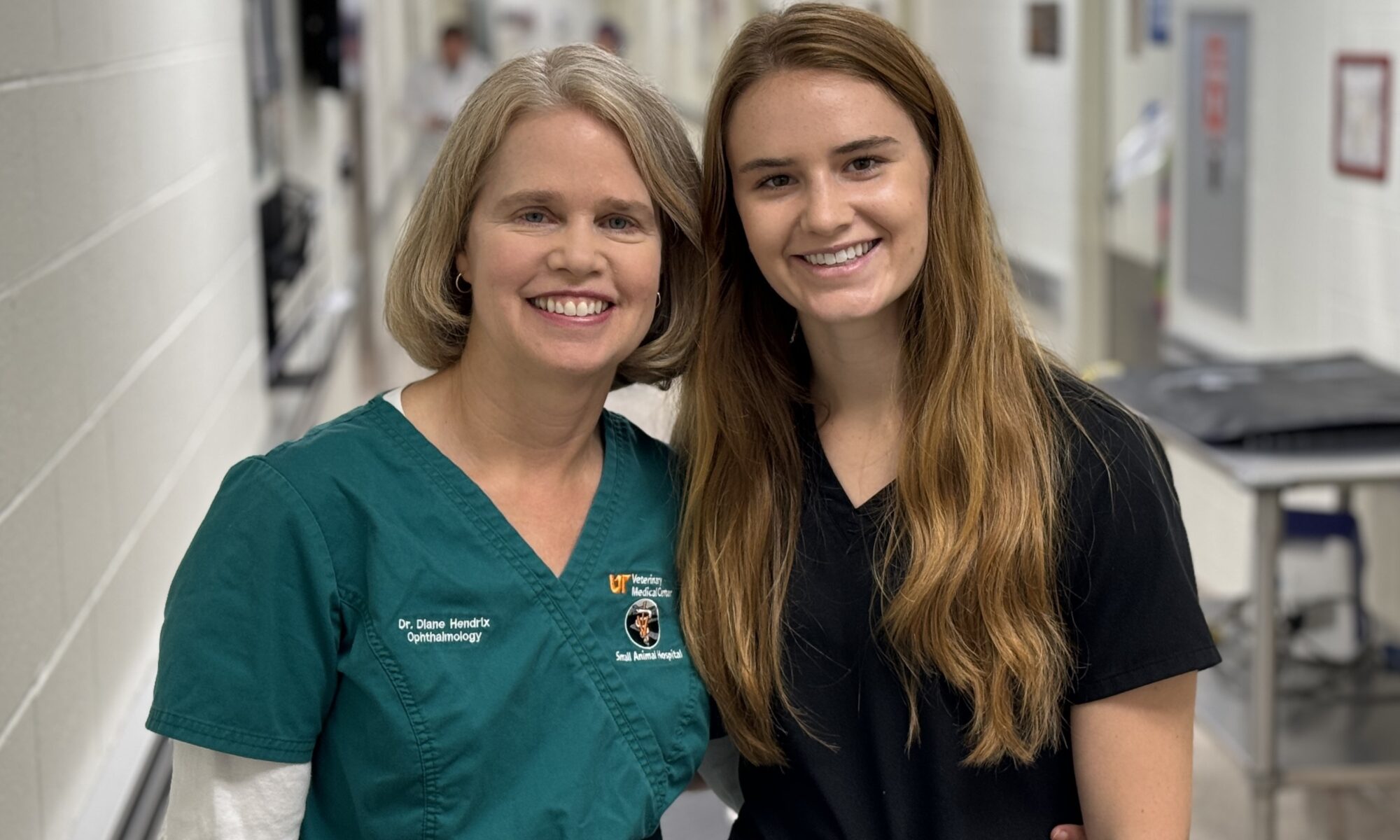 Dr. Diane Hendrix in screen scrubs stands next to her daughter, Emma, who is wearing a black scrub top. They are in a veterinary hospital hallway.