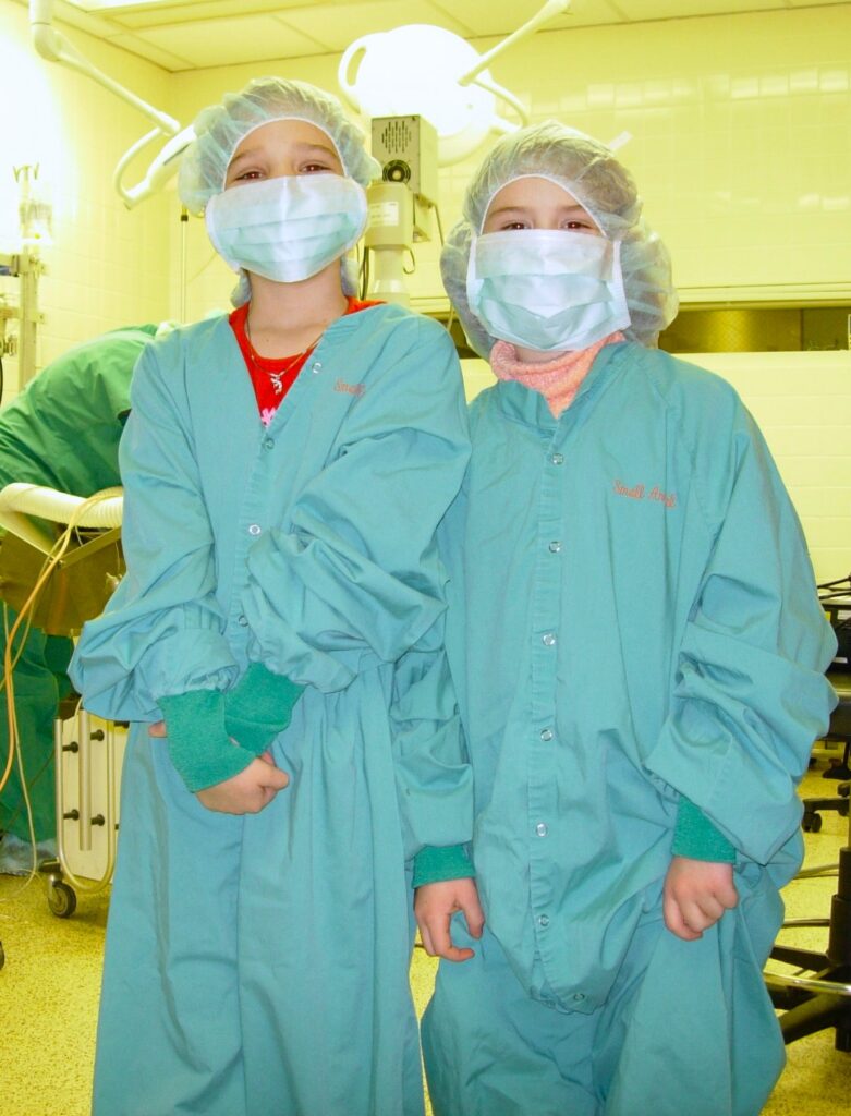 Two young girls wear oversized scrub suit, facemask and surgical bonnets. 