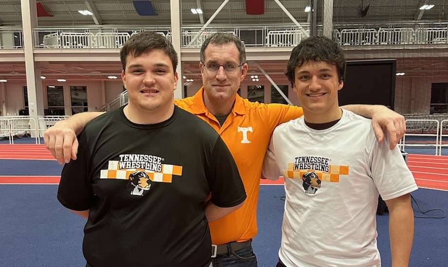 Wrestling coach wearing an orange shirt stands between two male wrestlers who have qualified for national tournament