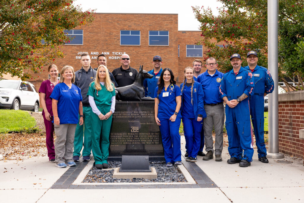 Group photo of 13 people in scrubs, flight gear, and police uniforms stand beside the War Dog Memorial