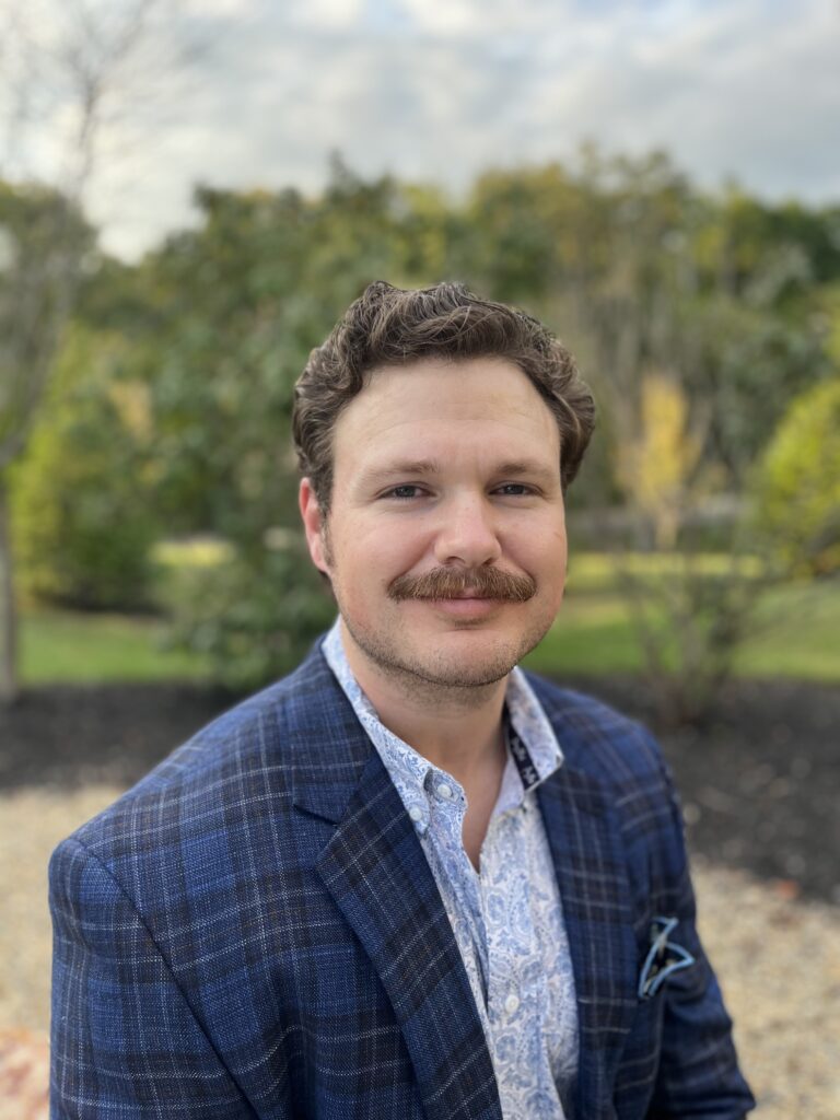 A man with dark hair and a mustache smiles at the camera while wearing a blue plaid jacket with trees in the background.