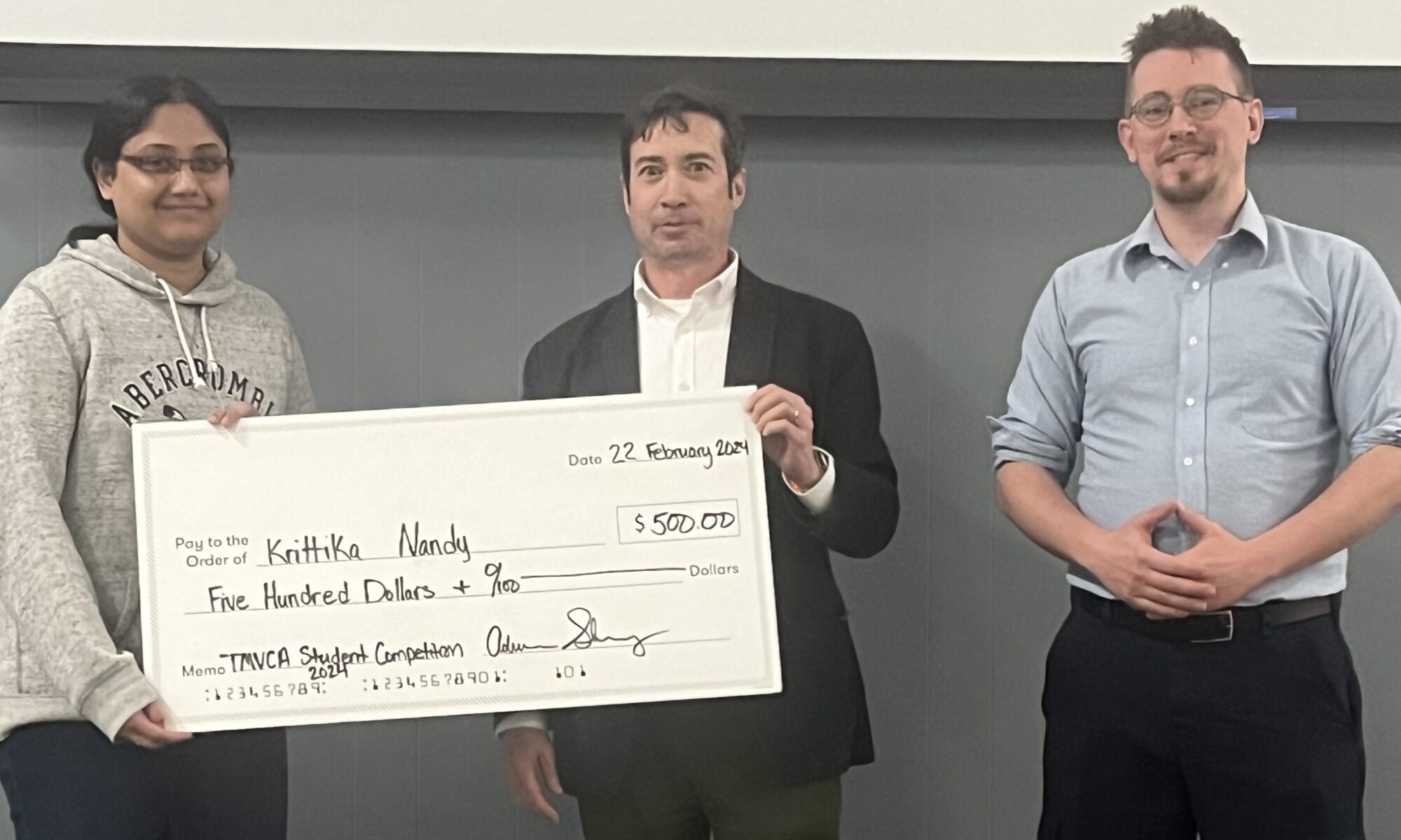 A female graduate student receives an oversized check for winning an oral presentation. Two men are also in the photo