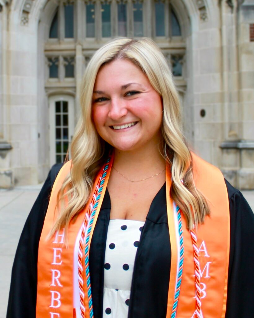 Mackenzie Furches is pictured wearing her graduation gown with an orange stole with Herbert written on it. She is standing in front of a building with a stone facade.