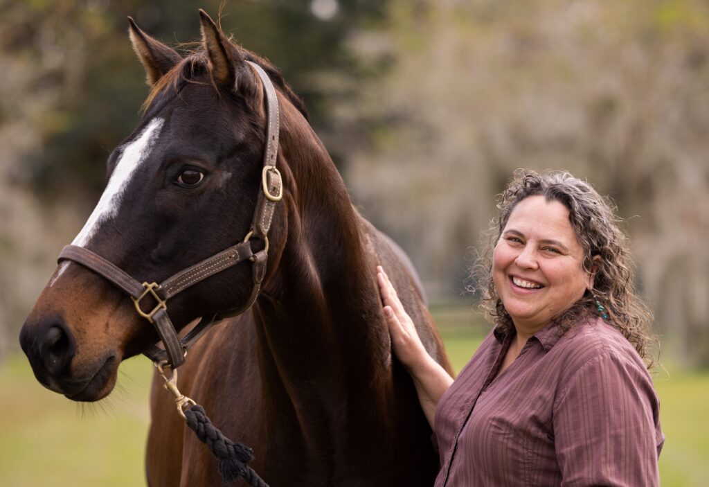 Dr. Martha Mallicote, wearing a plum-colored shirt stands next to a bay horse. 