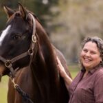 Dr. Martha Mallicote, wearing a plum-colored shirt stands next to a bay horse.