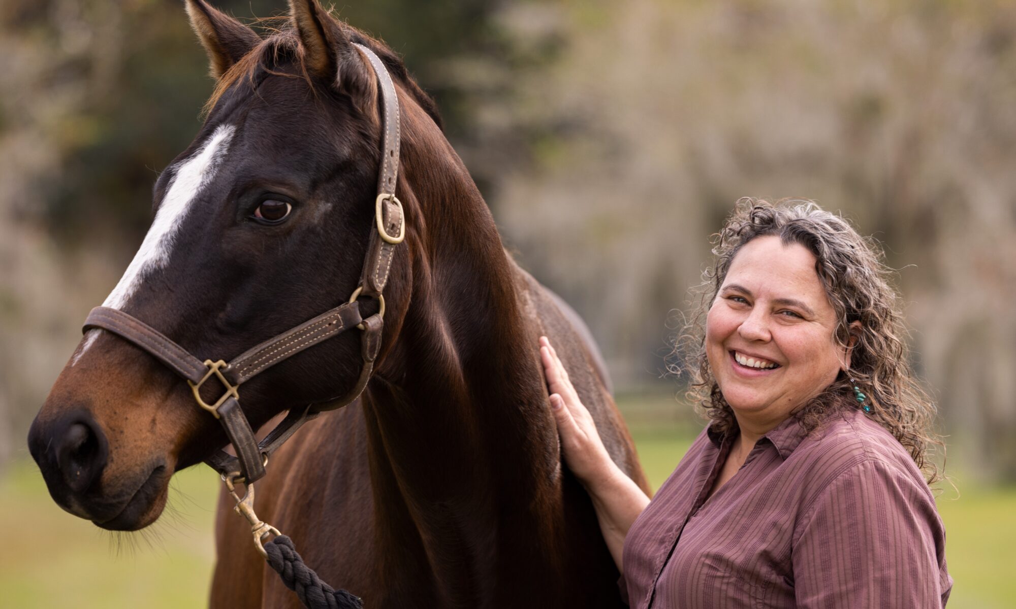 Dr. Martha Mallicote, wearing a plum-colored shirt stands next to a bay horse.