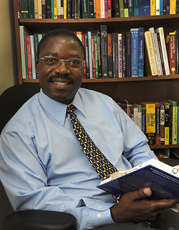 Dr. Agricola Odoi in light blue shirt reading a book in front of a bookcase