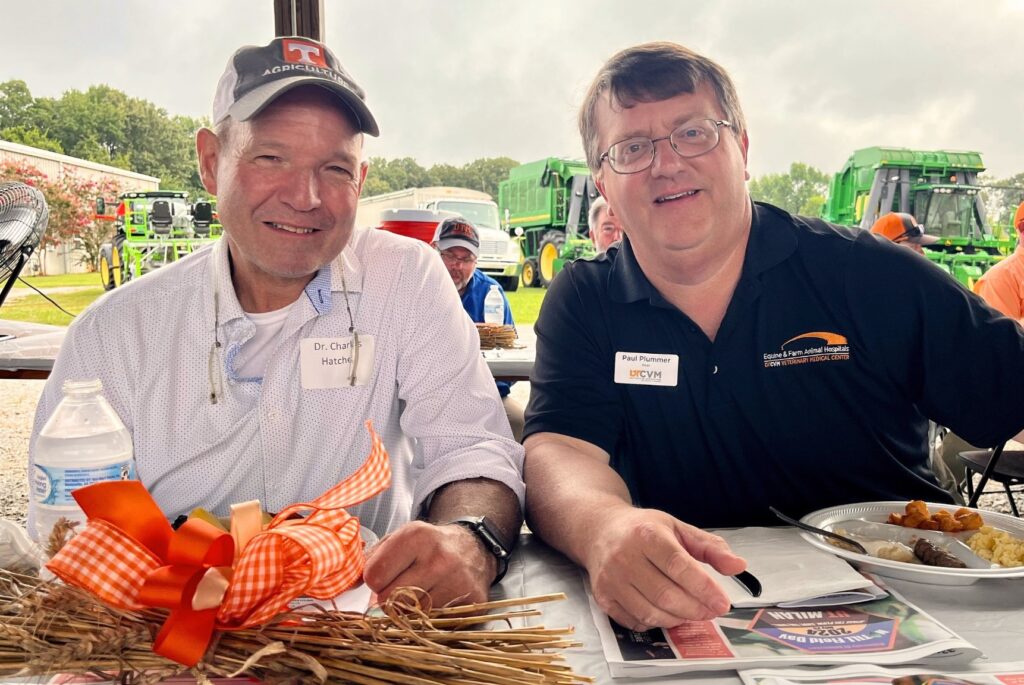 Charlie Hatcher in a baseball hat and white shirt is sitting with Paul Plummer who is wearing a black shirt. Both are looking across the table at the camera