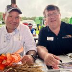 Charlie Hatcher in a baseball hat and white shirt is sitting with Paul Plummer who is wearing a black shirt. Both are looking across the table at the camera