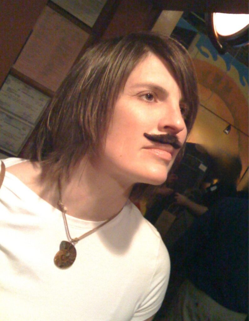 A woman wearing a white shirt looks wistfully toward the ceiling while sporting a face mustache. 