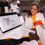 Professor Hameeda Sultana, wearing a white lab coat and orange gloves, describes a black and white image on a computer screen.