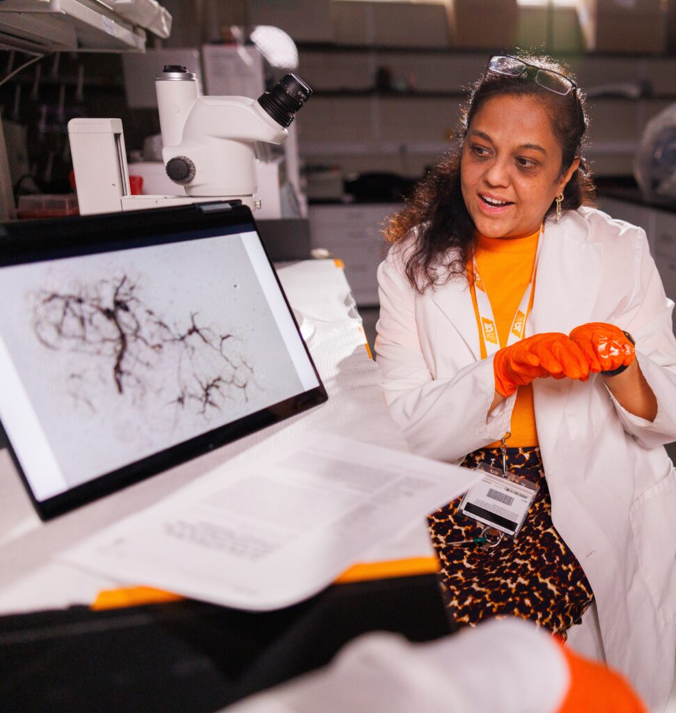 Professor Hameeda Sultana, wearing a white lab coat and orange gloves, describes a black and white image on a computer screen.