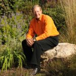 Dean Jim Thompson, wearing an orange shirt and black pants, sitting on a rock in the UT Trial Gardens