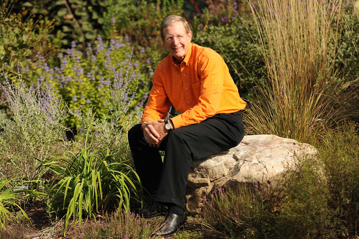 Dean Jim Thompson, wearing an orange shirt and black pants, sitting on a rock in the UT Trial Gardens