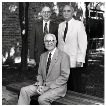 Three men pose for a photo, with one sitting and two standing behind him. 